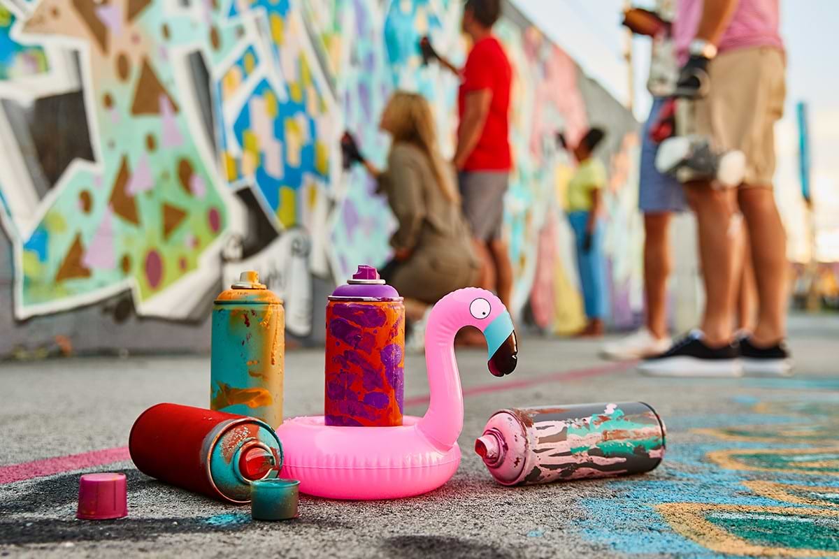 People painting colorful graffitis on the walls and a plastic pink flaming with paint' sprays on the floor